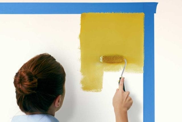 女人用滚轴刷和黄色油漆在墙上的一个区域，已经标记出用胶带屏蔽-油漆床头板- Goodhomesmagazine.com