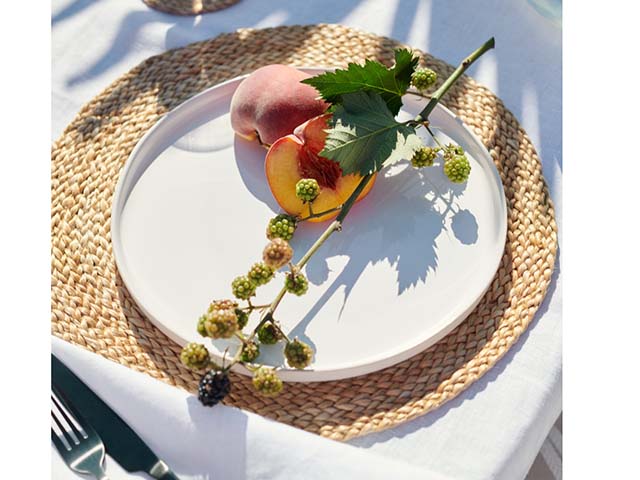 白色亚麻桌布上的圆形黄麻餐垫，上面有桃红和叶子，网址是goodhomesmagazine.com